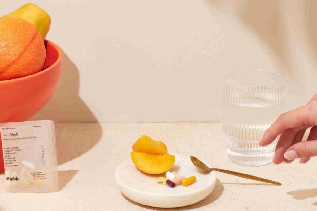 a bowl of fruit, a glass of water and a selection of Vitable vitamins, against a white backdrop